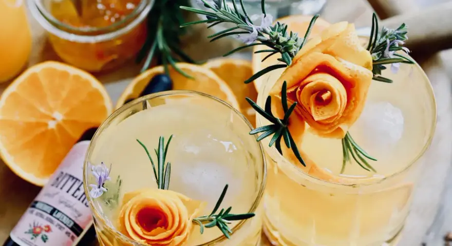 Zwei erfrischende Cocktails mit Orangenscheiben, Rosmarin und einer Orangenschalenrose. Daneben eine Flasche "Bitters" mit Lavendelgeschmack.