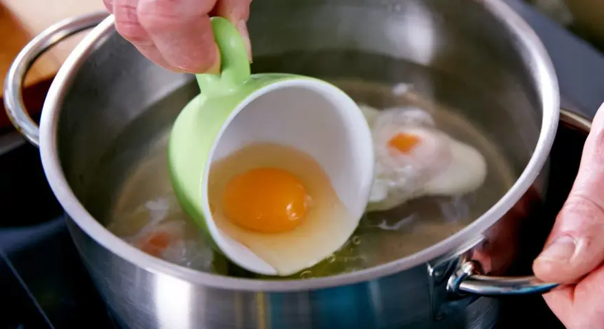 Ein rohes Ei wird aus einer grünen Tasse in kochendes Wasser gegossen, um verlorene Eier zuzubereiten.