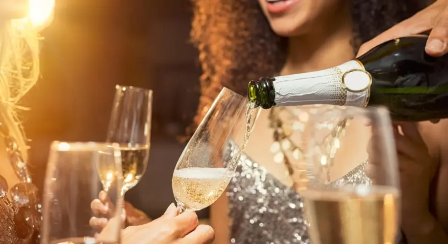 Sekt wird in ein Glas gegossen, während Freunde anstoßen. Der Unterschied zwischen Wein und Sekt ist die Kohlensäure, die Sekt prickelnd macht.