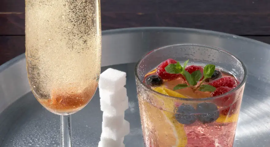 Sektglas mit sprudelndem Champagner-Cocktail, daneben ein Glas mit Beeren, Zitrone und Minze. Ein Stapel Zuckerwürfel.