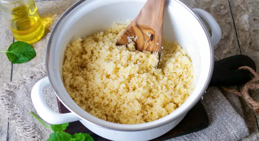 Frisch zubereiteter Couscous in einem Topf, bereit zum Servieren. Ein Holzlöffel lockert die Körner auf. Daneben eine Flasche Öl und Minzblätter.
