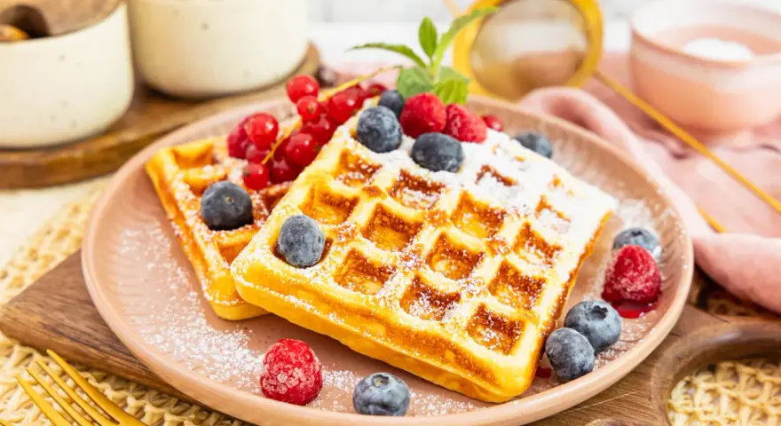 Zwei goldbraune Proteinwaffeln, bestäubt mit Puderzucker und garniert mit frischen Beeren und Johannisbeeren.