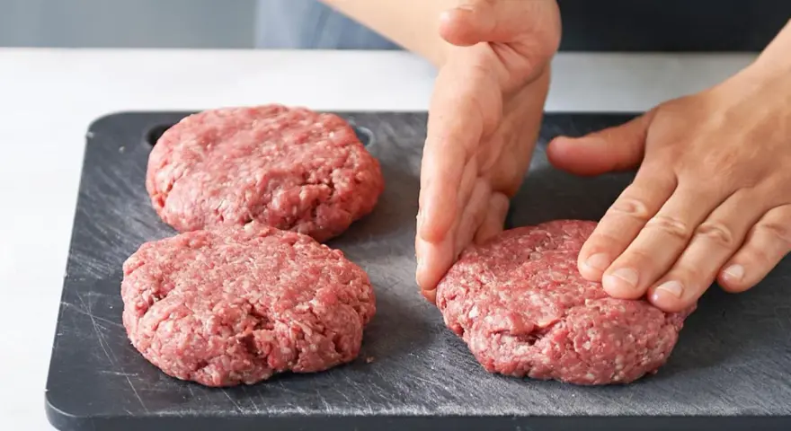 Hände formen rohe Burger-Patties auf einem dunklen Schneidebrett.