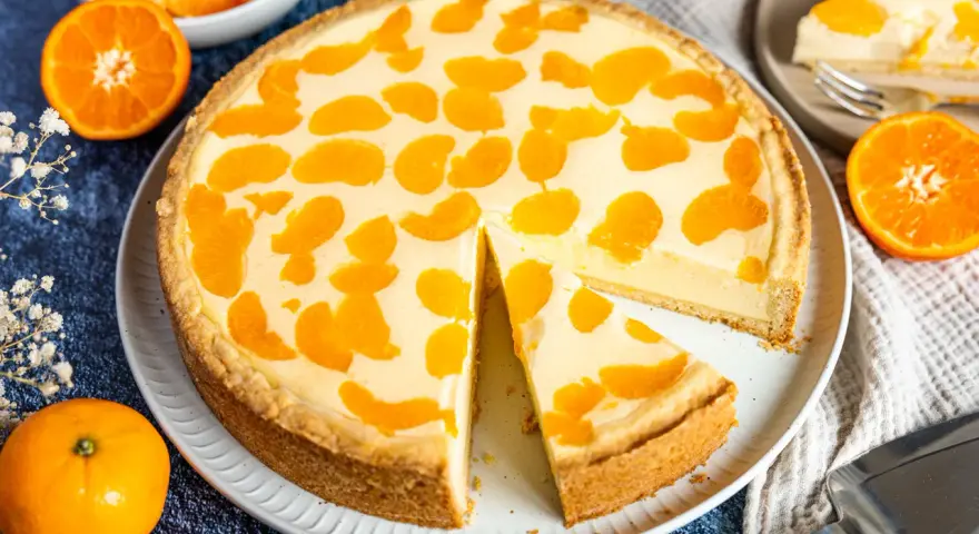 Ein saftiger faule weiber kuchen mit Mandarinenstücken, der auf einem Teller liegt. Daneben liegen frische Mandarinen und eine Tortenschaufel.