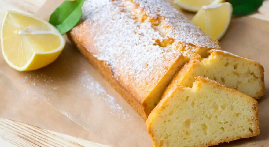 Saftiger veganer Zitronenkuchen, bestäubt mit Puderzucker, neben frischen Zitronenspalten und grünen Blättern.