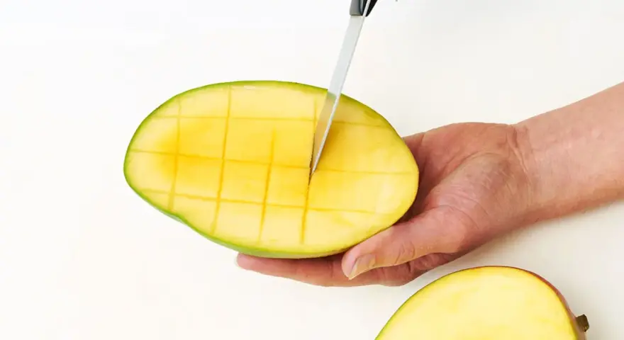 Eine Hand schneidet mit einem Messer eine reife Mango in Würfel. So einfach lässt sich die Frucht für den Verzehr vorbereiten.