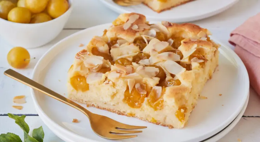 Ein Stück saftiger Mirabellenkuchen mit Mandelblättchen, serviert auf einem Teller mit einer goldenen Gabel. Daneben eine Schale voller frischer Mirabellen.