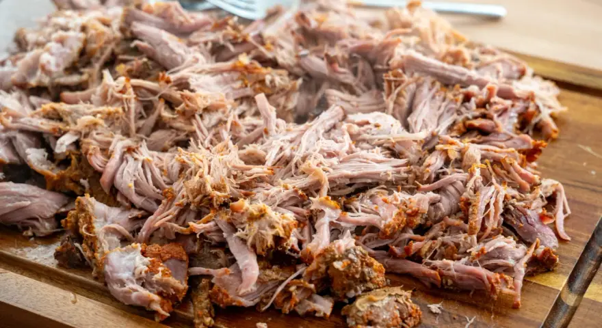 Saftiges pulled pork, zart zerfallen, liegt auf einem Holzbrett. Eine Gabel liegt bereit, um das Fleisch zu servieren.