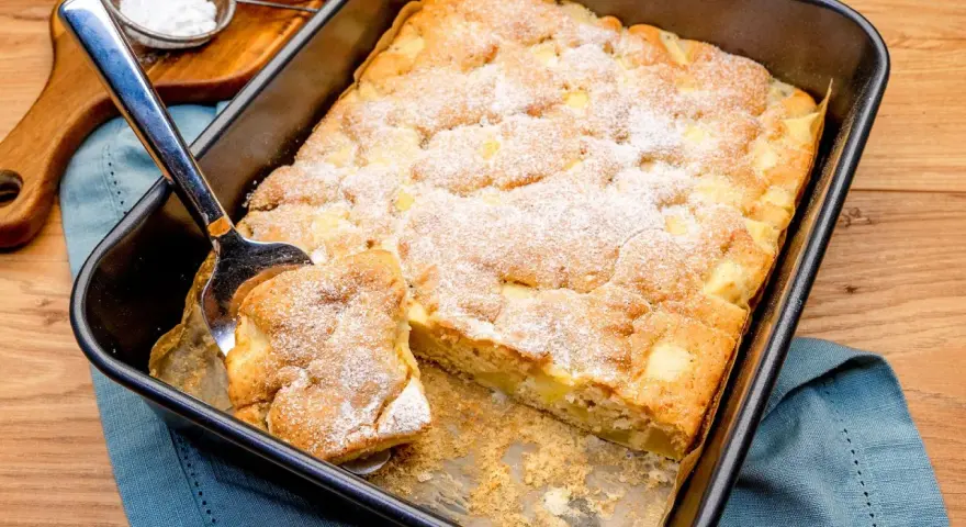Ein Stück vom schneller saftiger apfelkuchen vom blech wird mit einem Kuchenheber herausgehoben. Mit Puderzucker bestäubt.