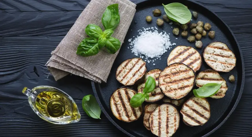 Gegrillte Auberginenscheiben mit Basilikum, Kapern und grobem Salz auf schwarzem Holz. Perfekt zum Aubergine grillen.