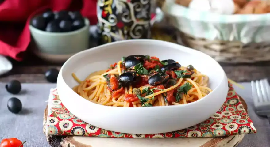 Eine Schüssel Spaghetti Puttanesca, garniert mit schwarzen Oliven und Kräutern, auf einem rustikalen Holzständer.