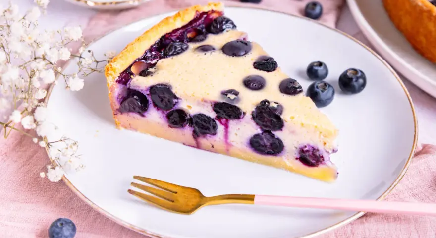 Ein Stück saftiger Heidelbeerkuchen Landfrauenart auf einem Teller, garniert mit frischen Beeren und einer Gabel.