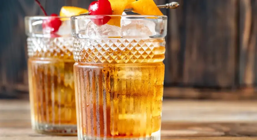 Zwei klassische Gin Old Fashioned Cocktails, garniert mit Kirschen und Orangenzesten, auf einem Holztisch.