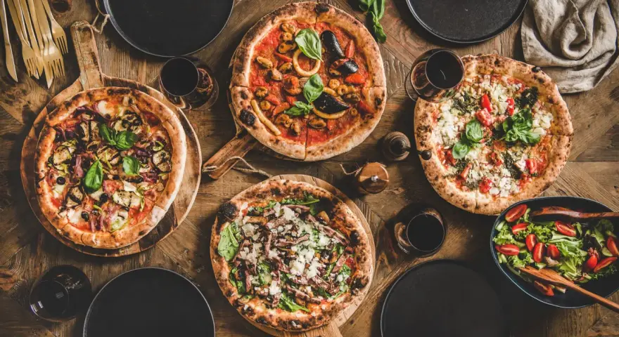 Vier leckere Pizzen und ein Salat auf Holztisch. Die Frage welcher Wein zu Pizza passt, wird hier mit Rotwein beantwortet.