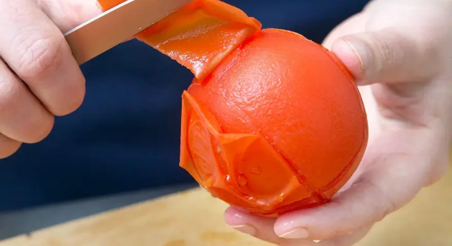 Hände ziehen mit einem Messer die Haut von einer Tomate ab. Die Tomate ist fast komplett geschält.