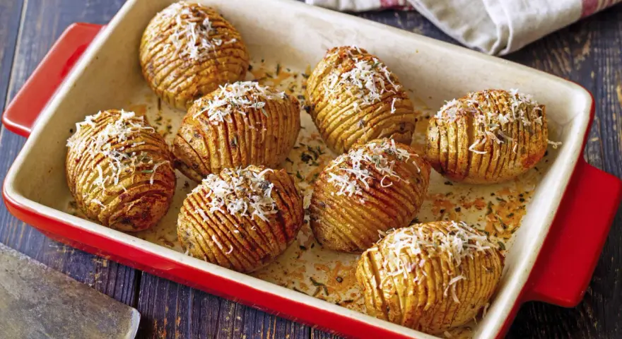 A red baking dish holds golden-brown hasselback kartoffeln, sprinkled with grated cheese and herbs.