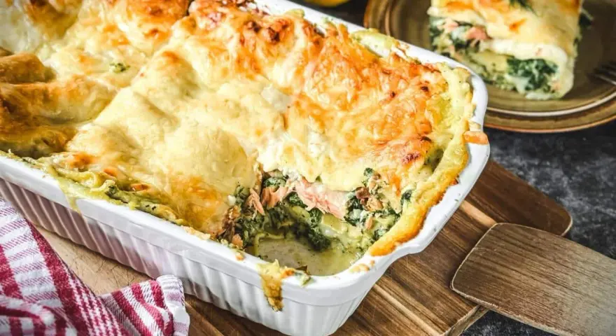Herzhafte Lachs Spinat Lasagne, goldbraun überbacken, dampft in einer weißen Auflaufform. Ein Stück auf einem Teller zeigt die Schichten.