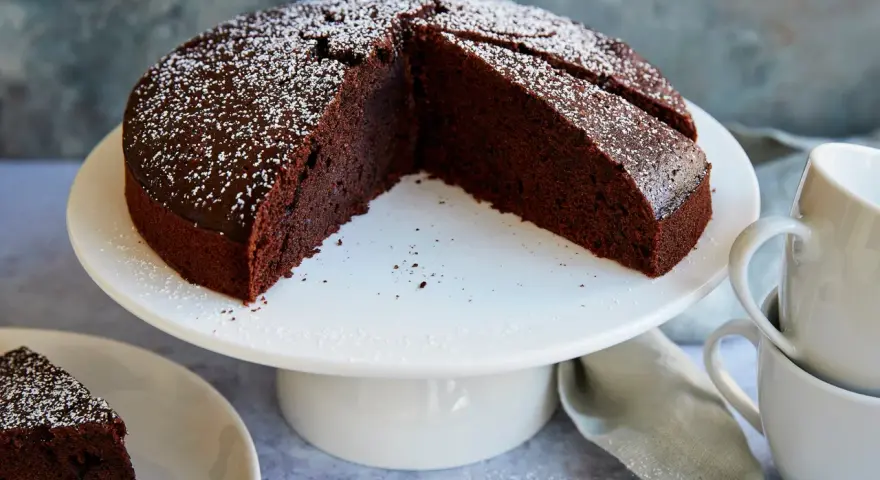 Ein saftiger veganer Schokokuchen, bestäubt mit Puderzucker, wartet auf Genuss. Daneben liegen zwei Tassen.