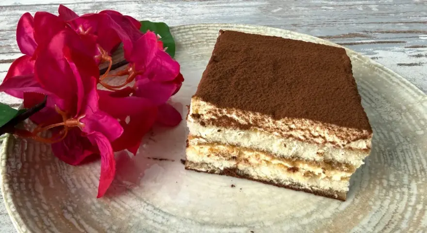 Ein Stück Tiramisu, perfekt für Ihr nächstes tiramisu rezept, liegt auf einem Teller neben leuchtend roten Blüten.