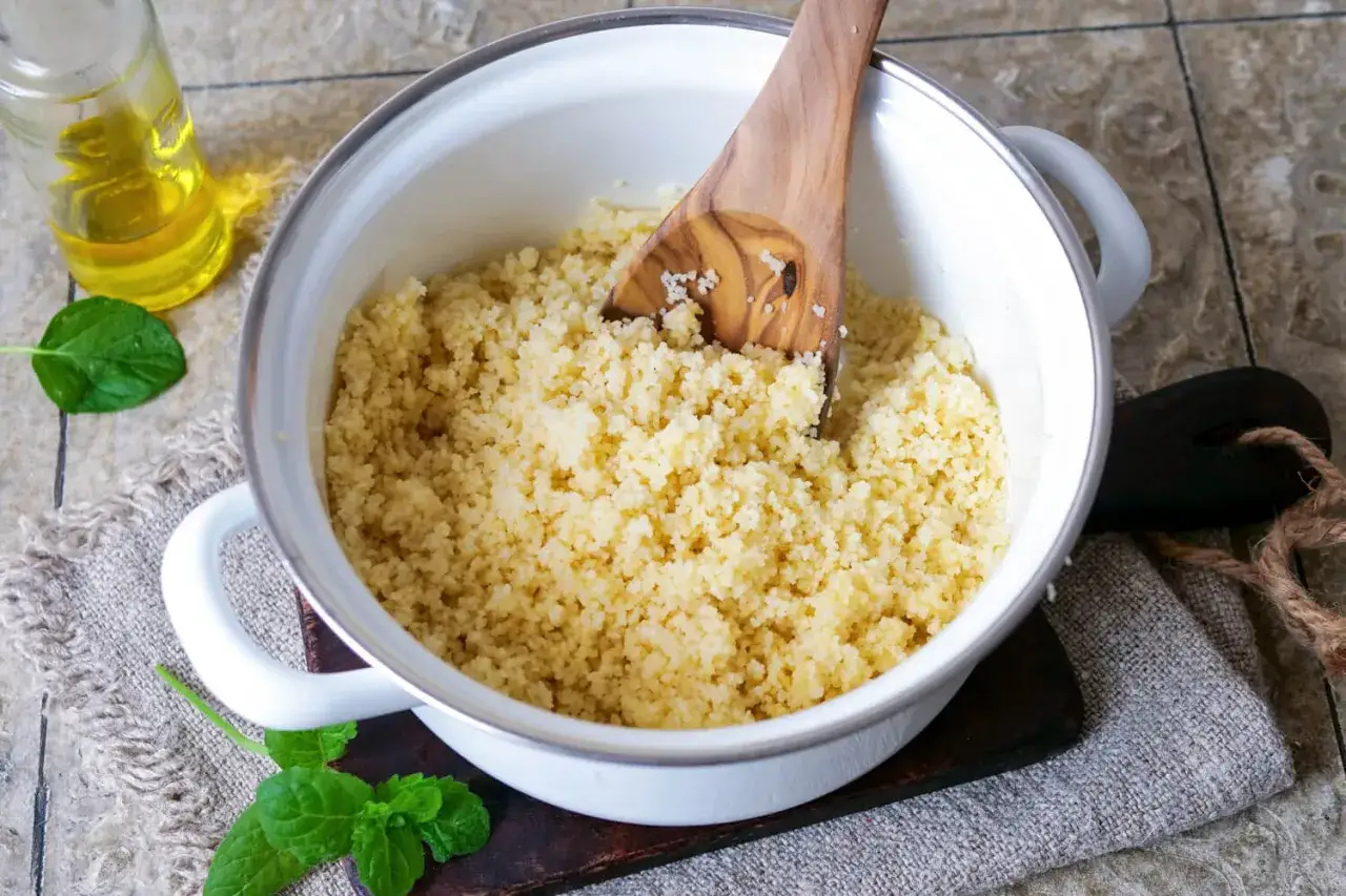 Frisch zubereiteter Couscous in einem Topf, bereit zum Servieren. Ein Holzlöffel lockert die Körner auf. Daneben eine Flasche Öl und Minzblätter.