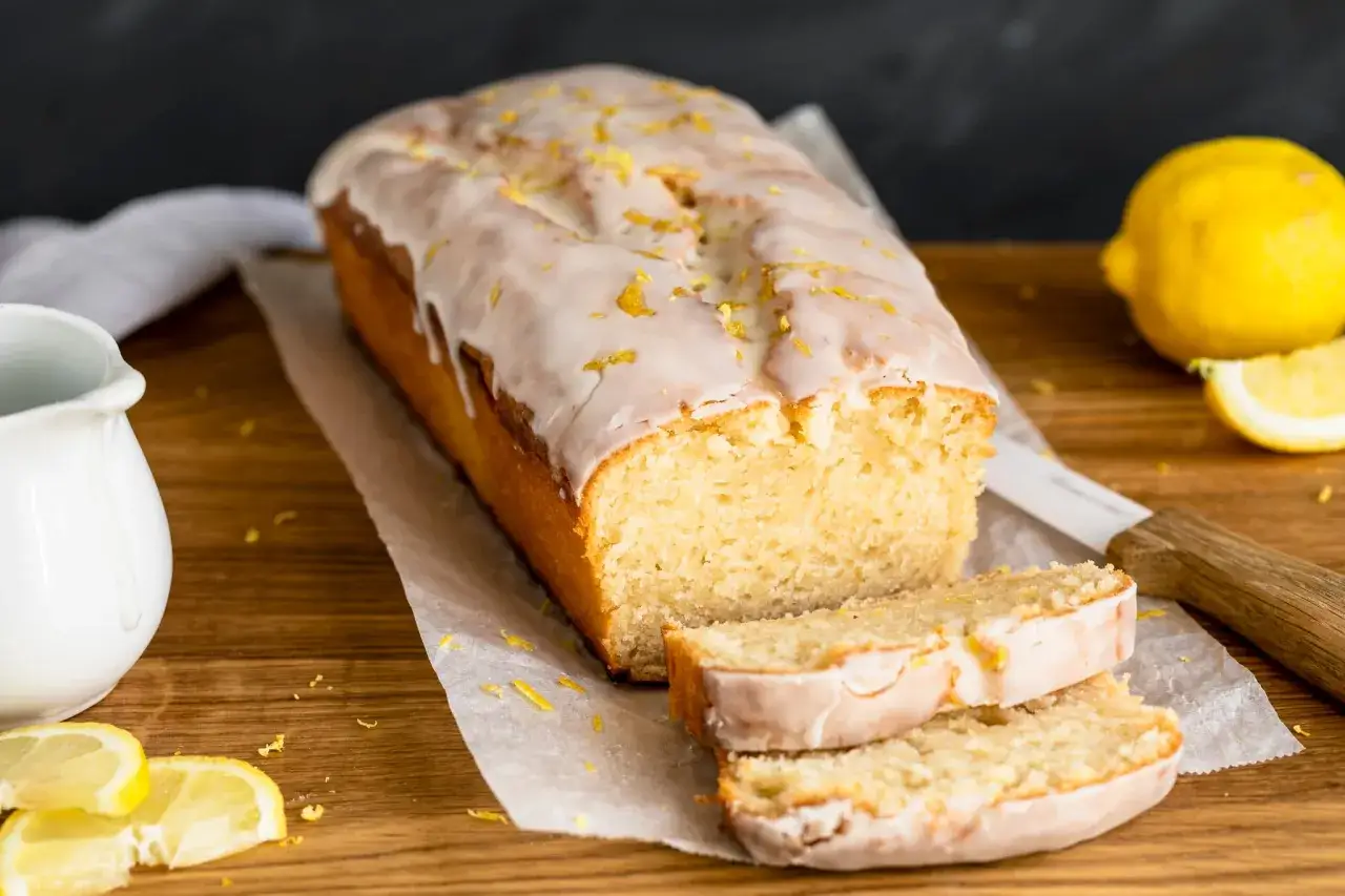 Ein saftiger, veganer Zitronenkuchen mit Zuckerguss und Zitronenschale bestreut, daneben frische Zitronenscheiben und ein Milchkännchen.
