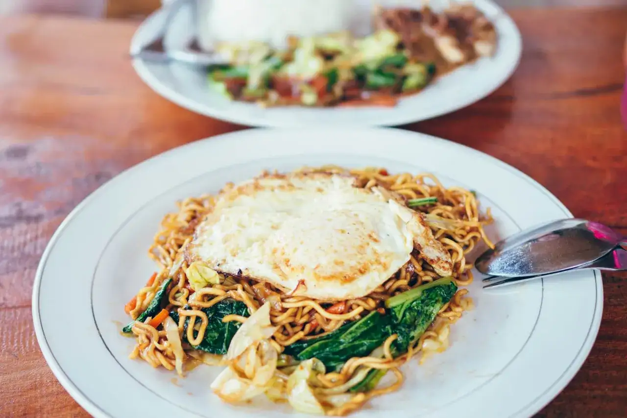Bami Goreng mit Spiegelei und Gemüse auf einem Teller, daneben ein Löffel.