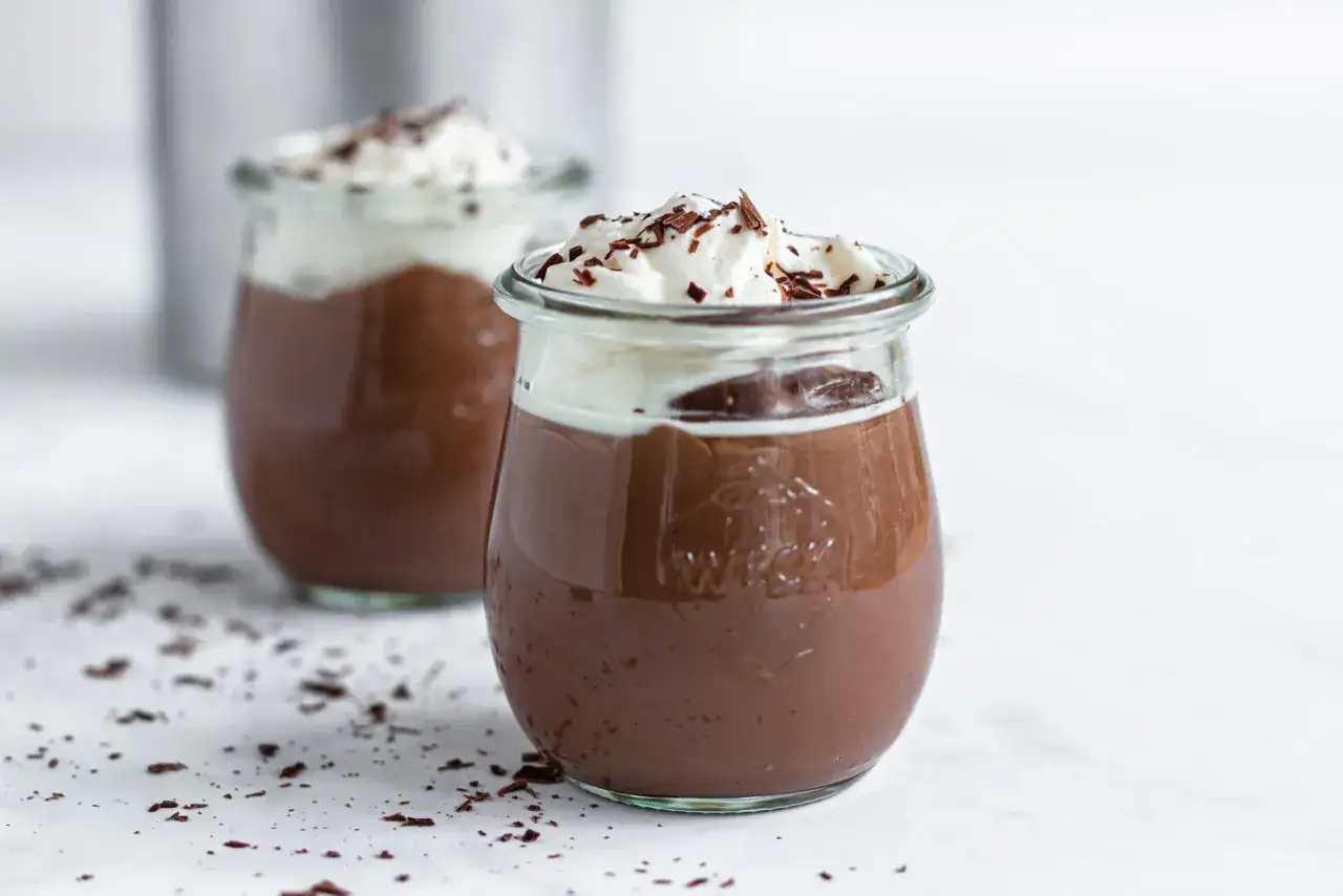 Zwei Gläser mit cremigem Schokopudding, gekrönt mit Schlagsahne und Schokostreuseln. Perfekt, um Schokopudding selber machen zu lernen!