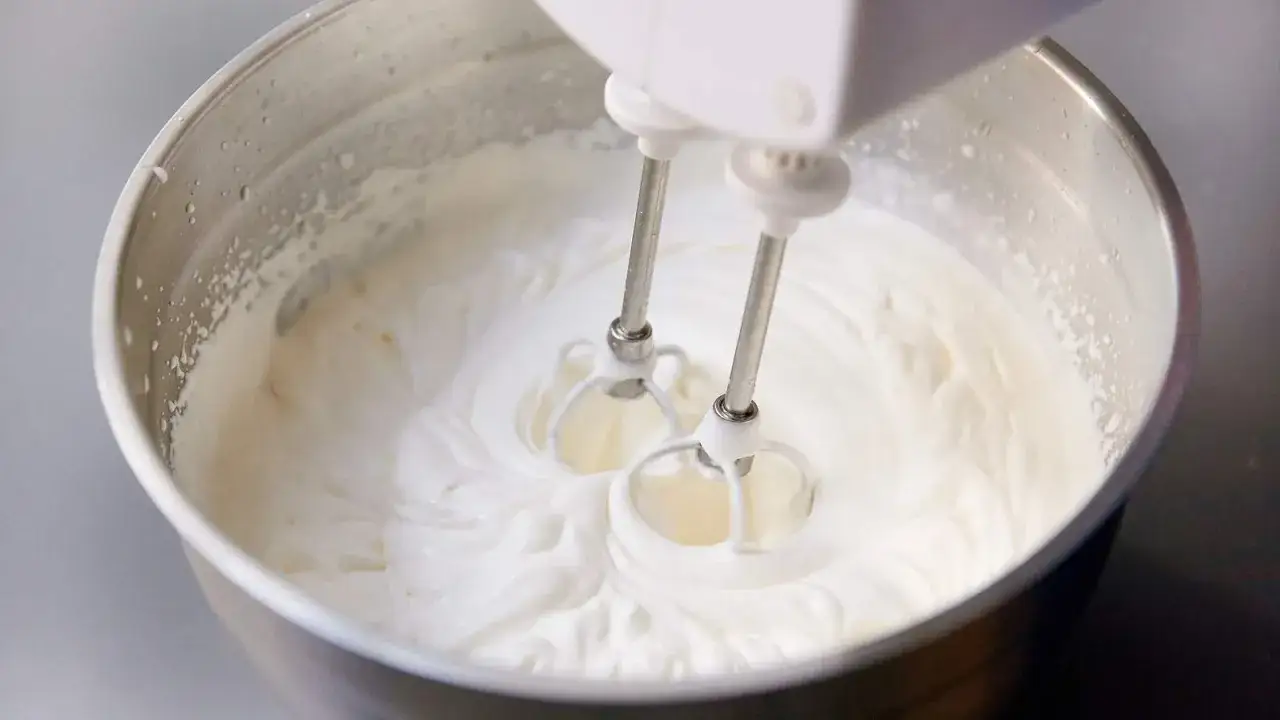 Ein Handmixer schlägt Sahne steif in einer Edelstahlschüssel. Die weiße, cremige Masse wirbelt um die Quirlen.