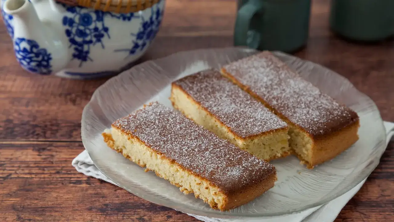 Drei rechteckige Stücke saftiger Mandelkuchen vom Blech, bestäubt mit Puderzucker, auf einem Glas-Teller.
