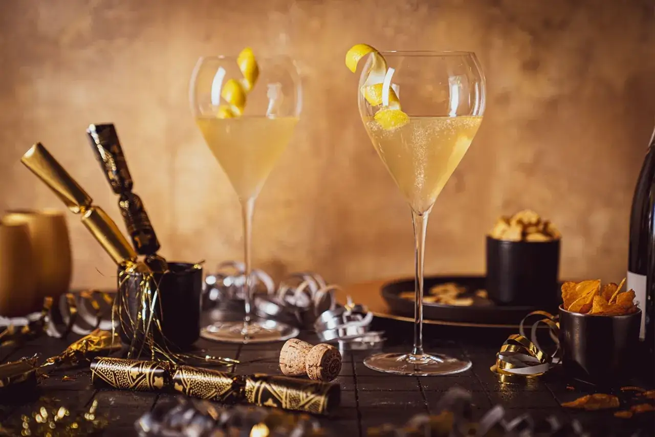 Zwei elegante Gläser mit einem prickelnden Champagner Cocktail, garniert mit Zitronenzesten. Festliche Knallbonbons und Konfetti deuten auf eine Feier hin.