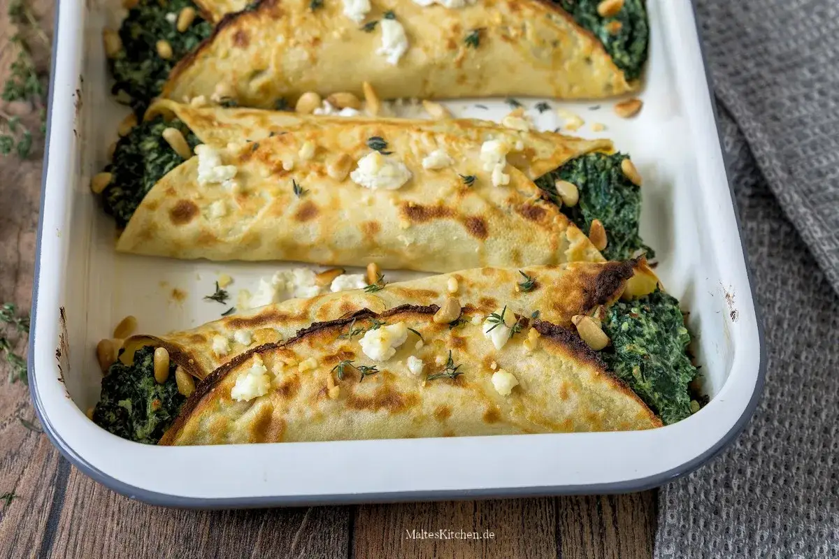 Herzhafte Pfannkuchen gefüllt mit Spinat und Feta, bestreut mit Pinienkernen und Thymian, in einer Auflaufform.
