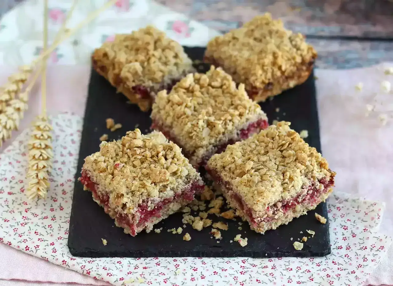 Vier Quadrate Himbeer Streuselkuchen auf einem schwarzen Brett, mit knusprigen Haferflocken-Streuseln und saftiger Himbeerfüllung.