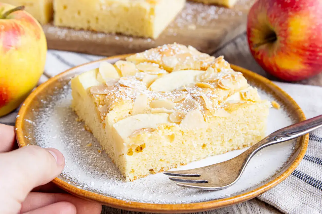 Ein Stück schneller saftiger Apfelkuchen vom Blech, bestreut mit Puderzucker und Mandelblättchen, daneben Äpfel.