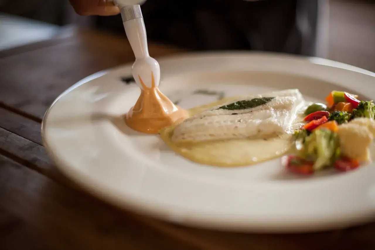 Ein Koch garniert ein Fischgericht mit einer cremigen Sauce. Was ist das? Espuma, eine leichte, luftige Zubereitung, wird hier auf den Teller gespritzt.