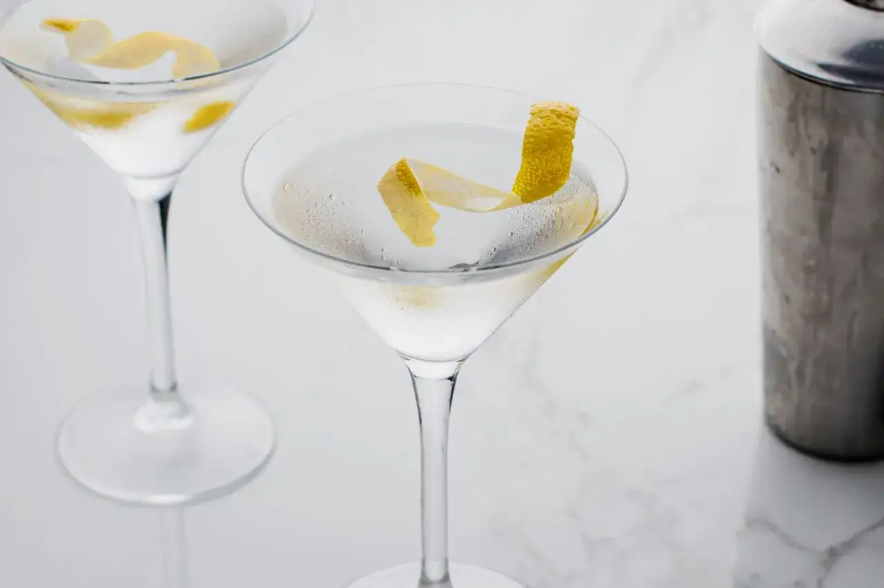Ein eisgekühlter Martini, wie ihn James Bond als Vesper genießt, mit Zitronenzeste garniert. Daneben ein Shaker.