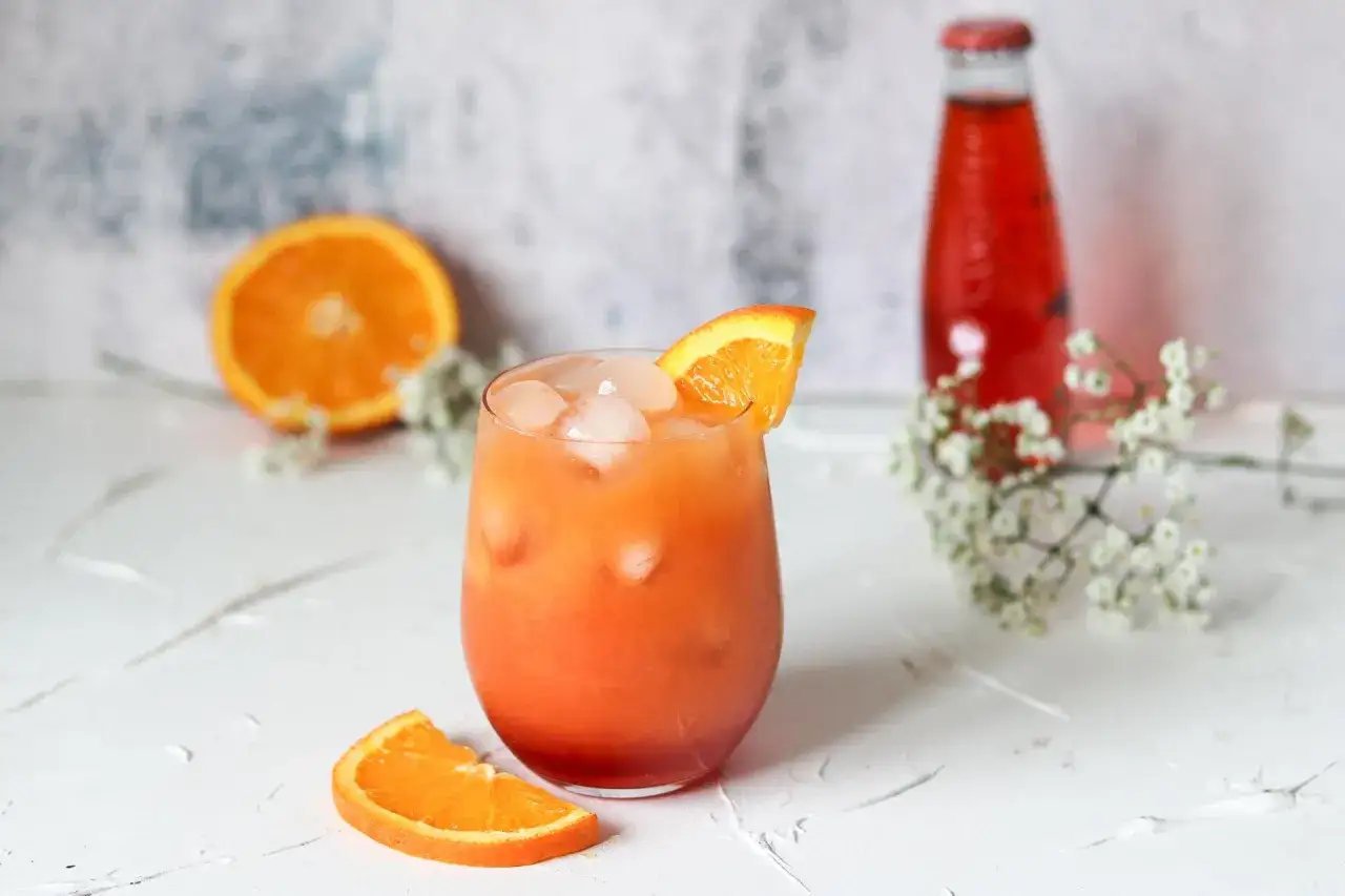Erfrischender Sanbitter Cocktail mit Eis und Orangenscheibe, daneben eine Flasche und frische Orangen.