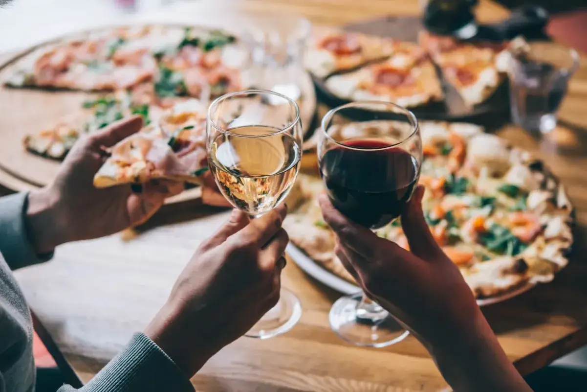 Zwei Hände halten Gläser mit Rot- und Weißwein, bereit für die Frage: welcher Wein zu Pizza? Mehrere Pizzen liegen auf dem Tisch.