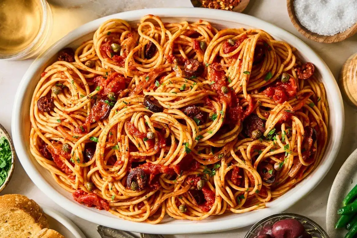 Ein Teller Spaghetti Puttanesca, garniert mit Petersilie, Oliven und Kapern. Daneben sind Brot und grüne Bohnen zu sehen.