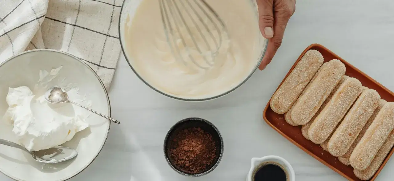 Zutaten für Tiramisu: Löffelbiskuits, Kakao, Kaffee und eine Schüssel mit geschlagener Sahne, die zeigt, welche Sahne zum Kochen am besten geeignet ist.