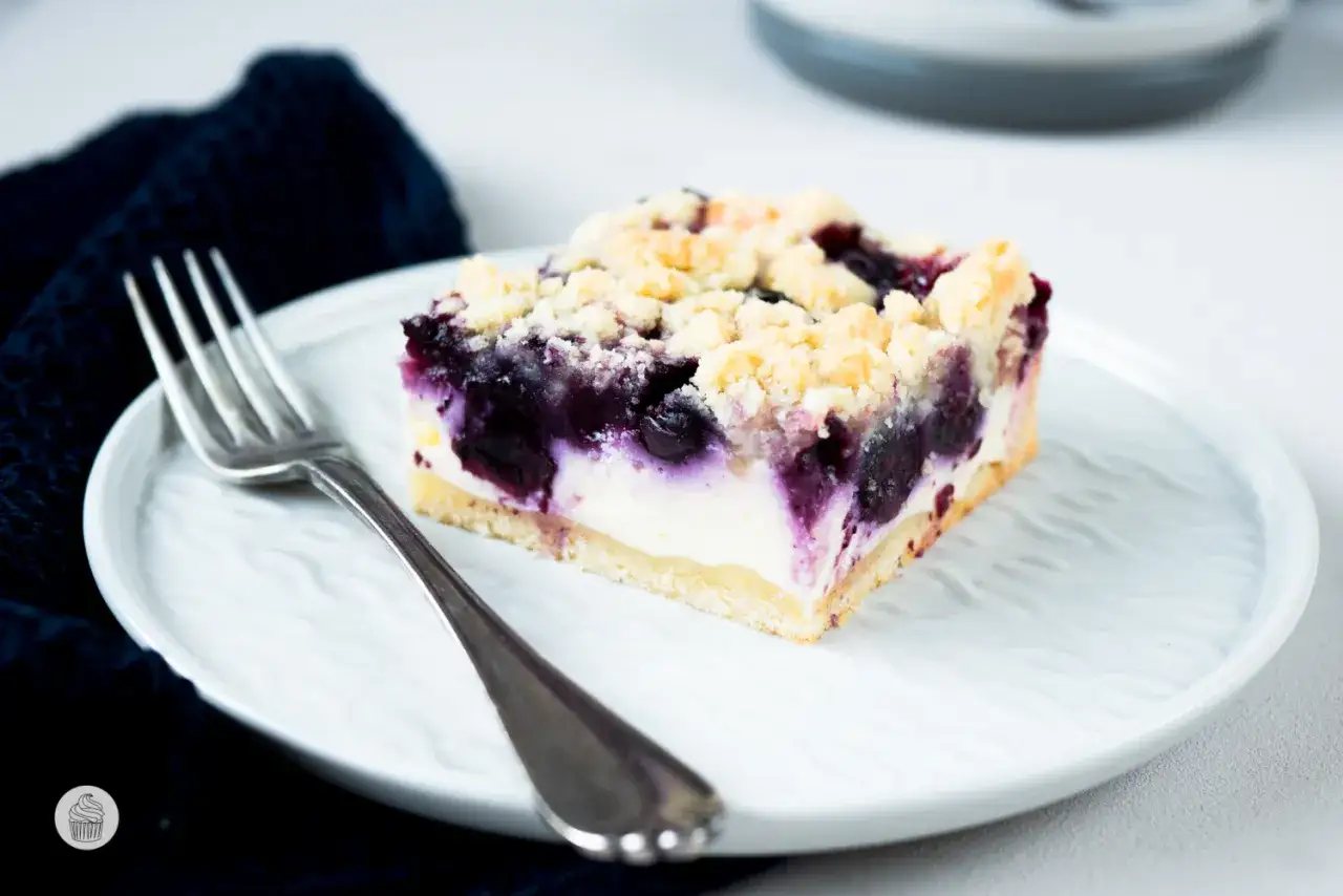 Ein Stück Heidelbeerkuchen Landfrauenart mit Streuseln und einer cremigen Füllung auf einem weißen Teller, daneben eine Gabel.