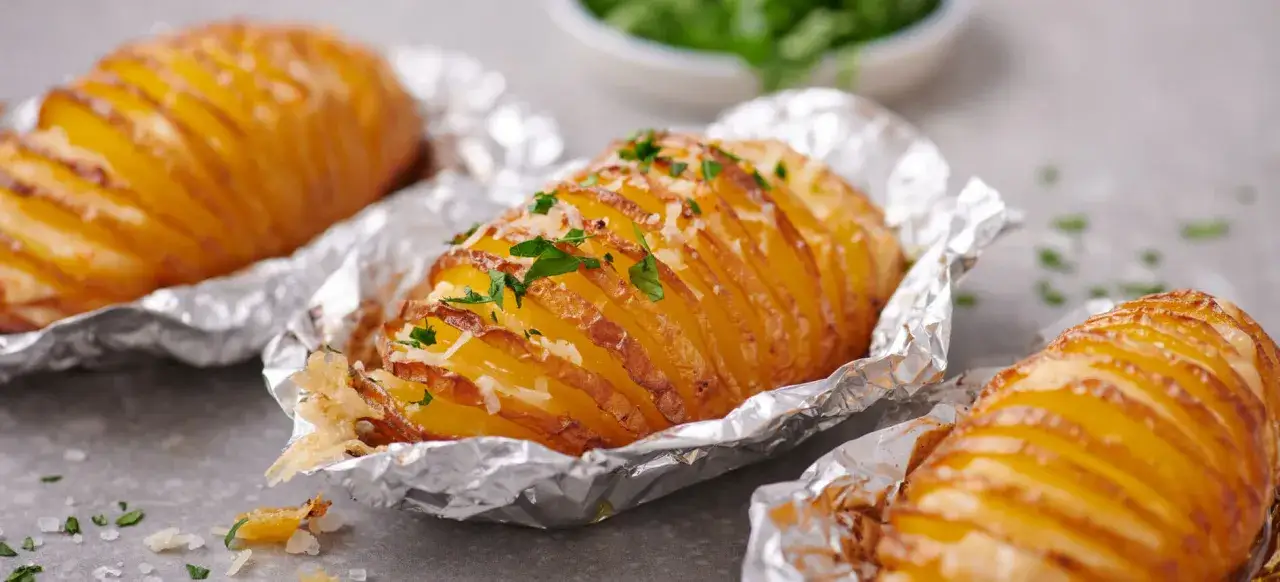 Drei goldbraune Hasselback Kartoffeln, in Alufolie gebacken und mit Käse und Petersilie bestreut, duften verlockend.