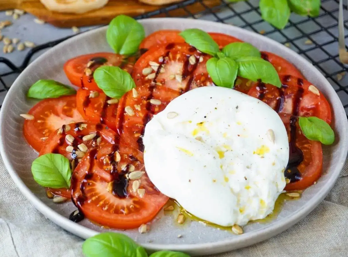 Ein Teller mit Tomatenscheiben, garniert mit Basilikum, Sonnenblumenkernen und Balsamico-Dressing. In der Mitte liegt ein Klacks cremige Burrata, die zeigt, was ist Burrata: ein frischer Mozzarella-Käse mit cremigem Kern.
