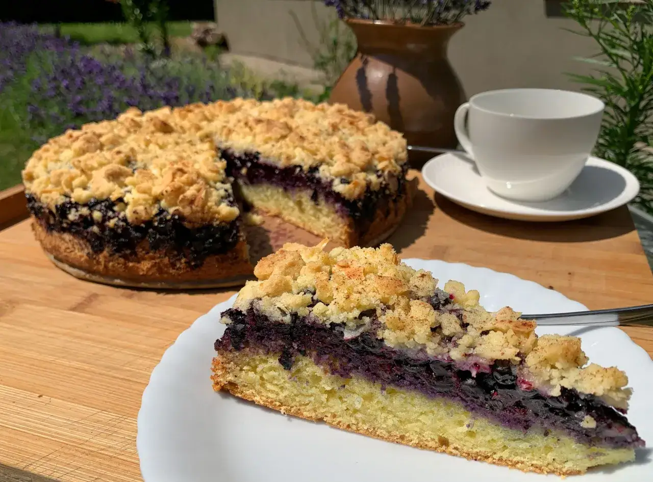 Ein saftiger Blaubeerkuchen mit Streuseln, ein Stück auf einem Teller, daneben eine Tasse Kaffee.