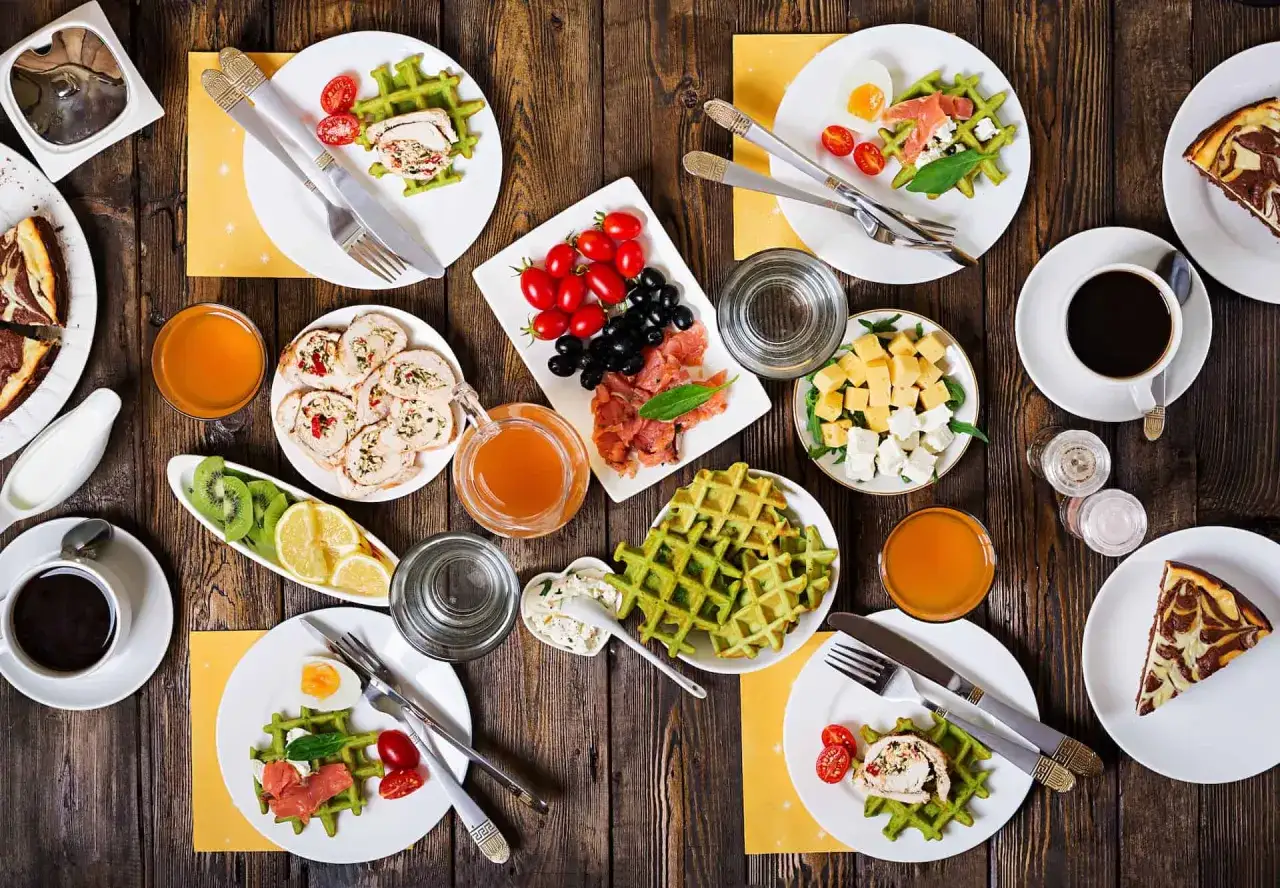 Osterbrunch Ideen: Grüne Waffeln mit Lachs, Röllchen, Käse, Obst und Kuchen auf Holztisch.
