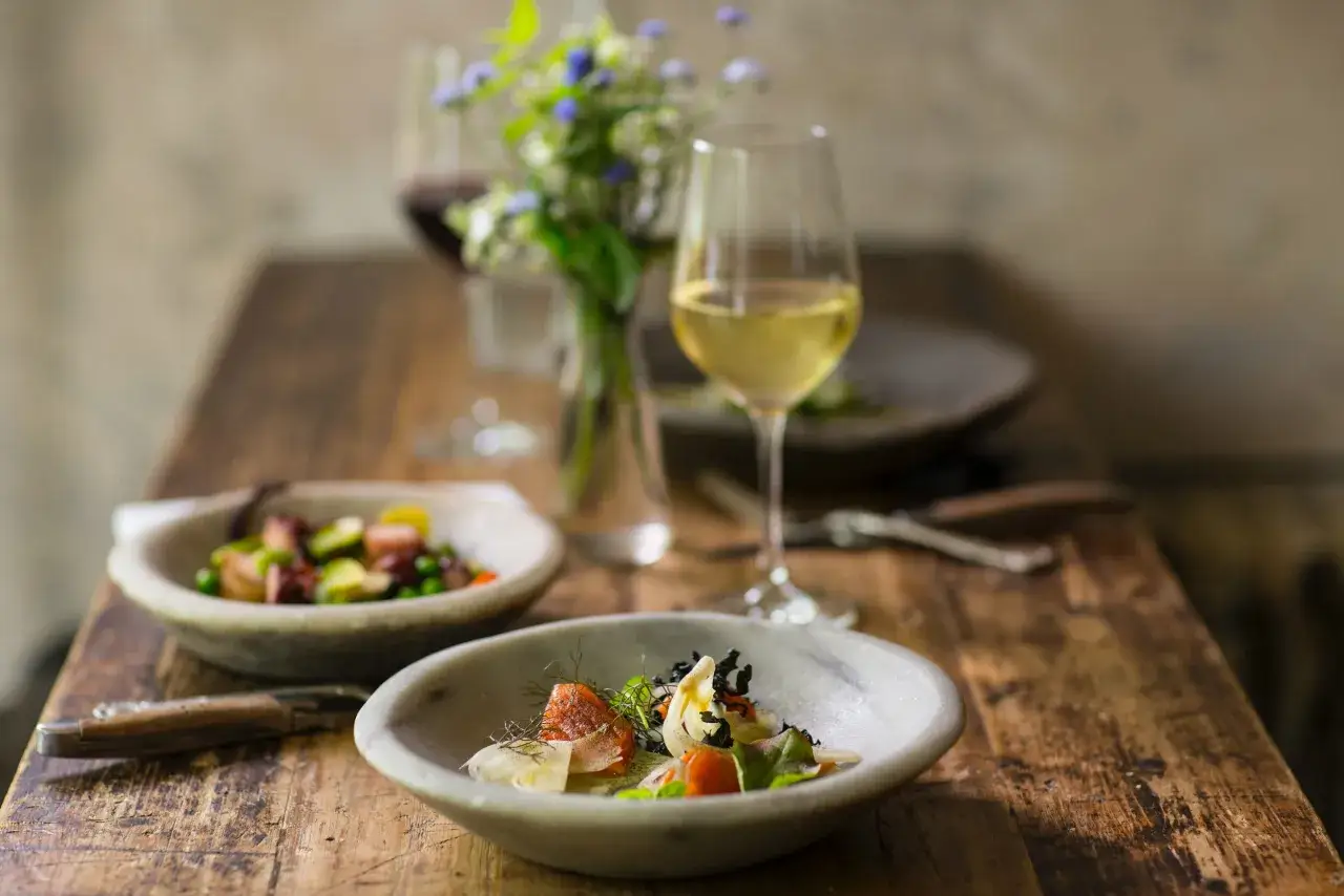 Ein gedeckter Tisch mit zwei Tellern, gefüllt mit appetitlichem Essen, und zwei Gläsern Wein. Ein kleiner Blumenstrauß in einer Vase sorgt für Frische.
