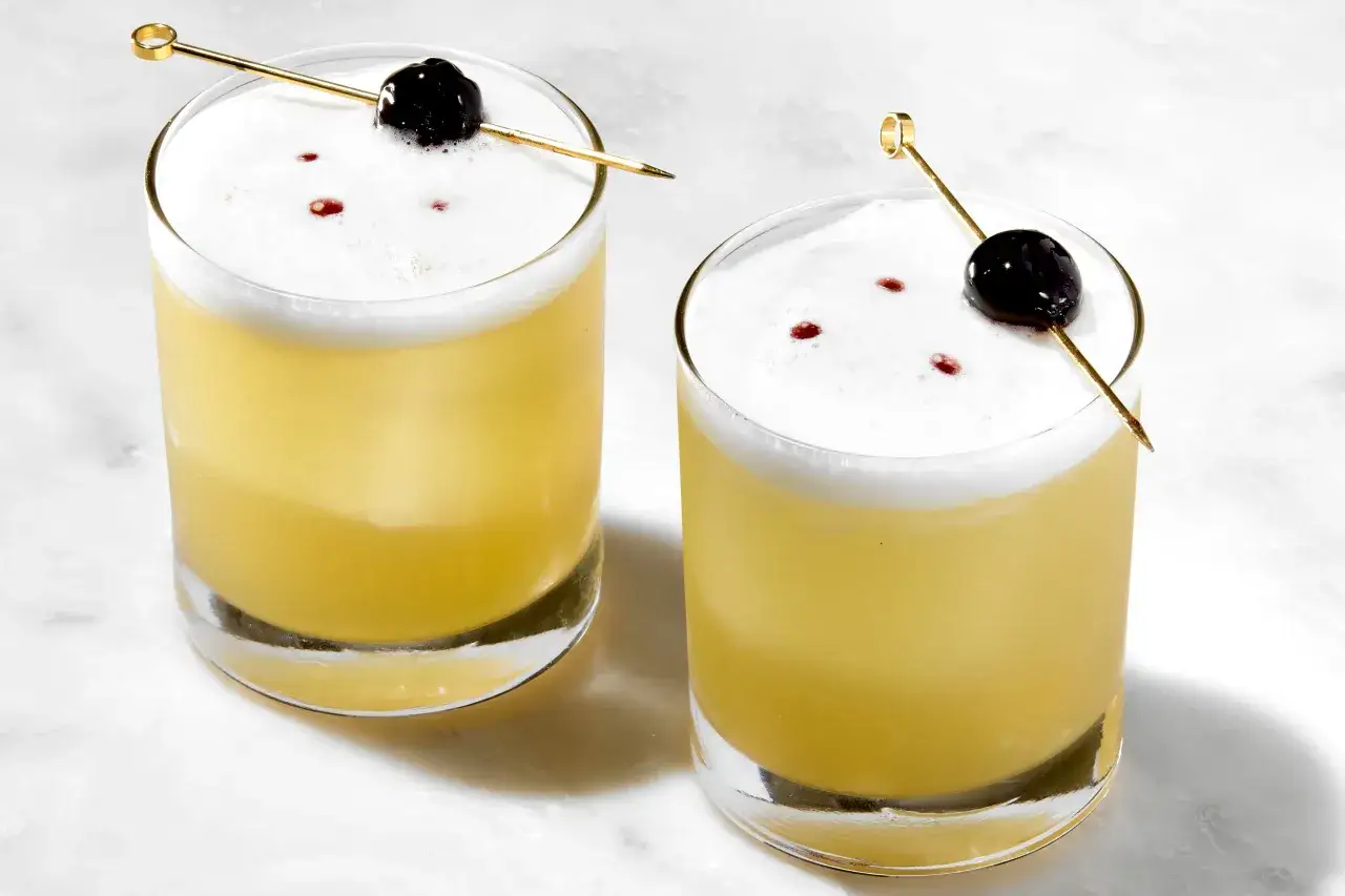 Zwei erfrischende Sour Cocktails mit Schaumhaube, Kirsche und einem Hauch von Bitter. Perfekt für ein Sour Cocktail Rezept.