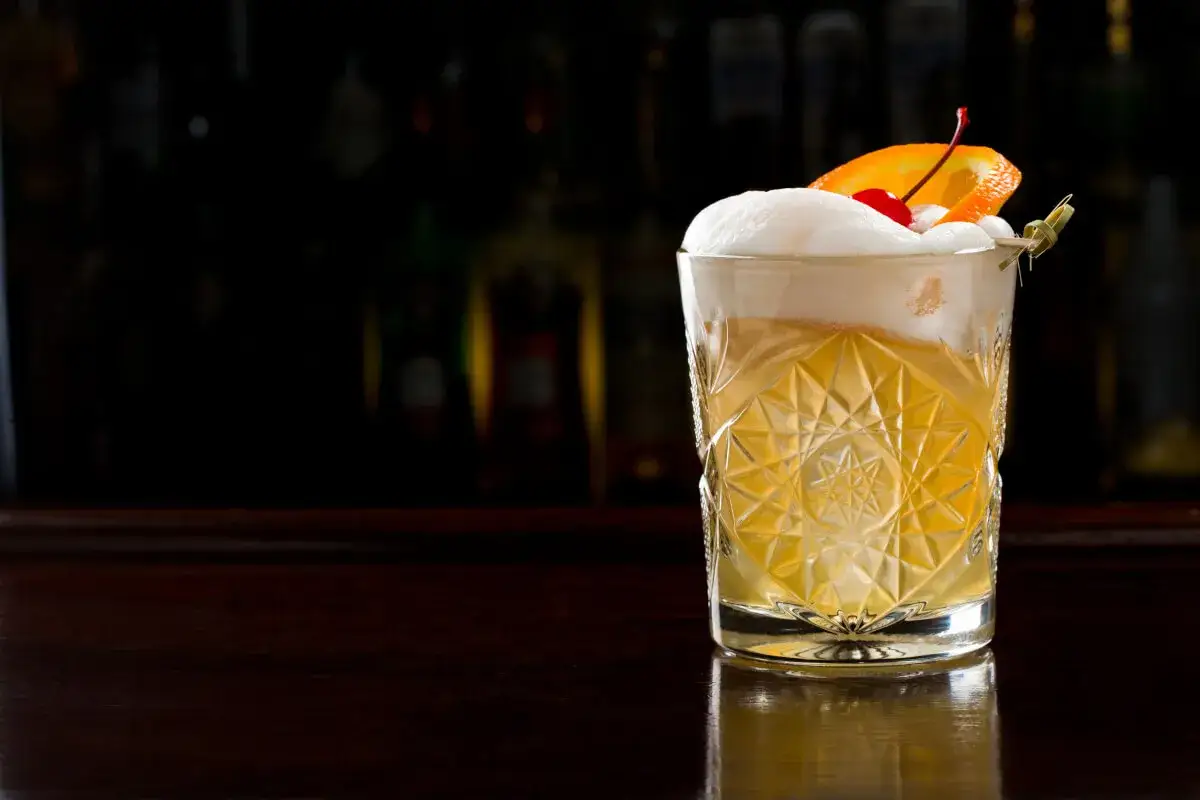 Ein Gin Old Fashioned mit Schaumhaube, Orangenscheibe und Kirsche auf einem dunklen Tresen.