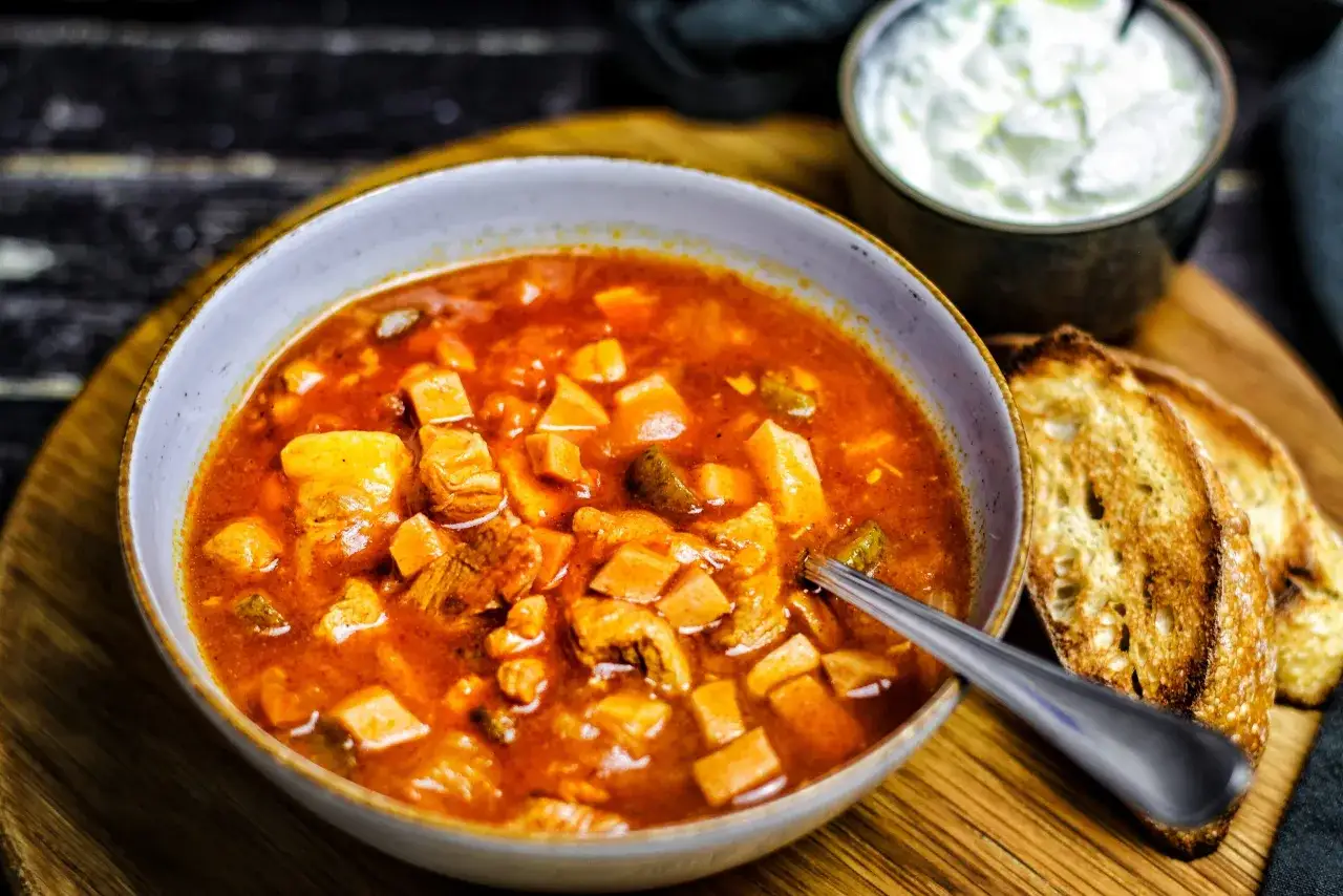 Herzhaftes Soljanka Rezept: Eine dampfende Sch&uuml;ssel voller Fleisch, Kartoffeln und Gew&uuml;rzen, serviert mit ger&ouml;stetem Brot und einem Klecks Sauerrahm.