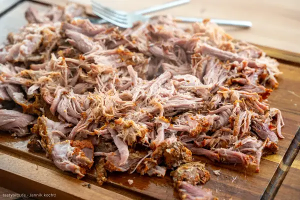 Saftiges pulled pork, zart zerfallen, liegt auf einem Holzbrett. Eine Gabel liegt bereit, um das Fleisch zu servieren.