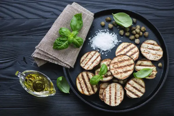 Gegrillte Auberginenscheiben mit Basilikum, Kapern und grobem Salz auf schwarzem Holz. Perfekt zum Aubergine grillen.